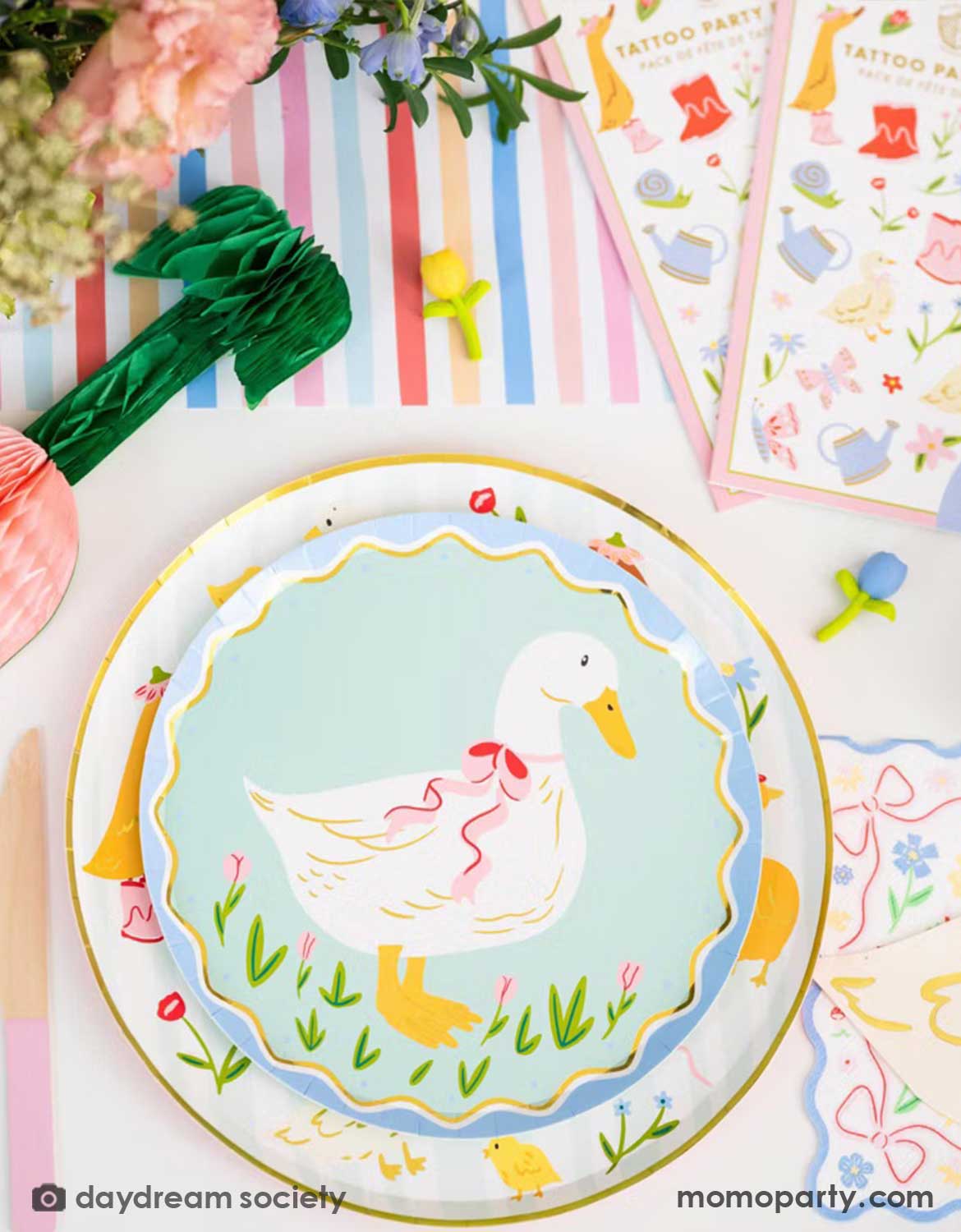 An overhead shot of a children's Easter table setting with colorful flowers, tulip honeycomb decorations, goose themed party plates, napkins, party cups, pink cutlery and multiple kid's Easter small gifts with bunny designs on a colorful striped runner. A perfect idea for an Easter celebration, a baby shower, kid's Silly Goose themed birthday party or a spring gathering.