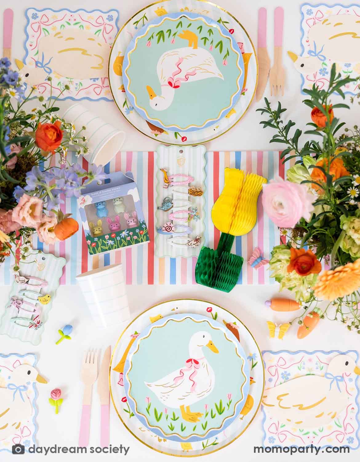 An overhead shot of a children's Easter table setting with colorful flowers, tulip honeycomb decorations, goose themed party plates, napkins, party cups, pink cutlery and multiple kid's Easter small gifts with bunny designs on a colorful striped runner. A perfect idea for an Easter celebration, a baby shower, kid's Silly Goose themed birthday party or a spring gathering.