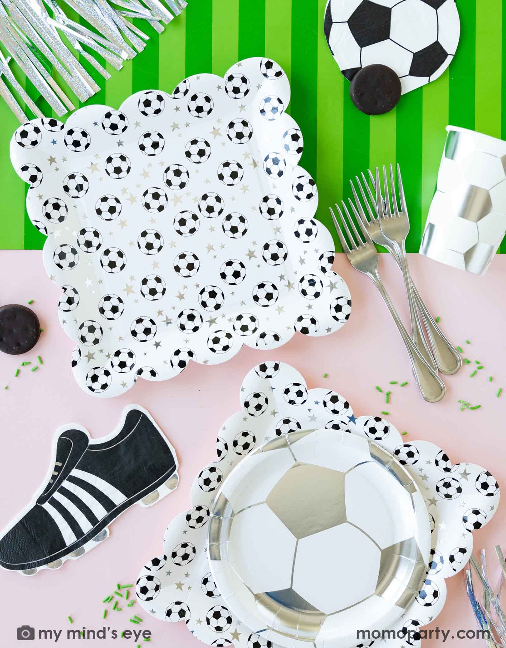 A chic soccer themed party table featuring Momo Party's soccer party collection including silver soccer shaped plate, party cup, soccer ball shaped napkin, soccer cleat shaped napkin and green striped soccer table runner on a light pink table. With some snacks, green sprinkles around, this makes a great inspo for a chic girly kid's soccer themed birthday party or a championship team celebration!