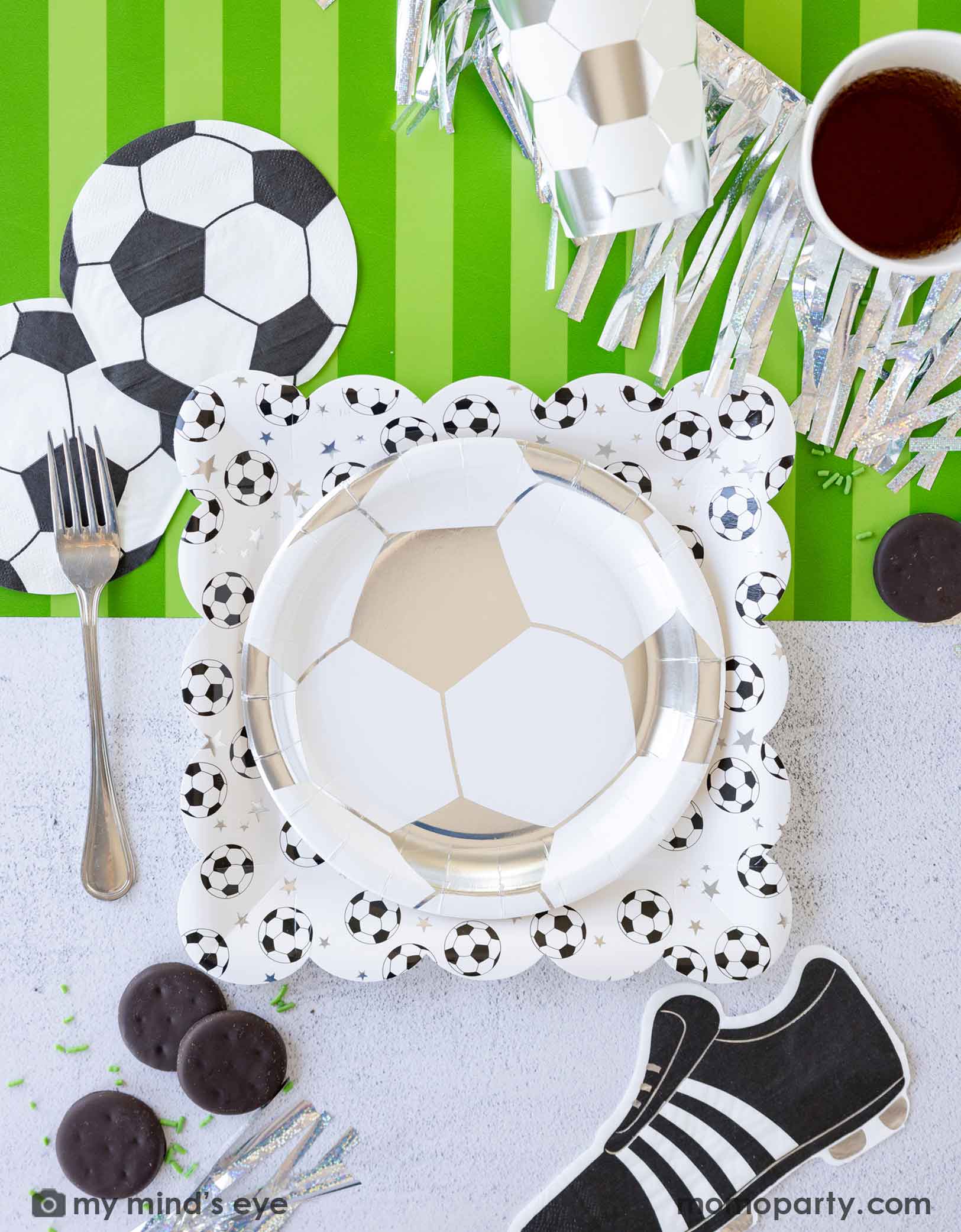 A fun soccer themed party table featuring Momo Party's soccer party collection including silver soccer shaped plate, party cups, soccer ball shaped napkins, soccer cleat shaped napkins and green striped soccer table runner by My Mind's Eye. With some snacks, green sprinkles around, this makes a great inspo for a fun kid's soccer themed birthday party or a championship celebration!