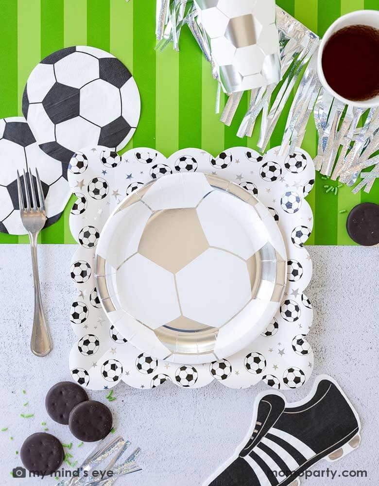 A fun soccer themed party table featuring Momo Party's soccer party collection including silver soccer shaped plates and party cups, soccer shaped napkins, cleat shaped napkins and soccer table runner by My Mind's Eye. With some snacks, silver streamers, green confetti and silver utensil around, this makes a great inspo for a fun kid's soccer themed birthday party or a championship celebration!