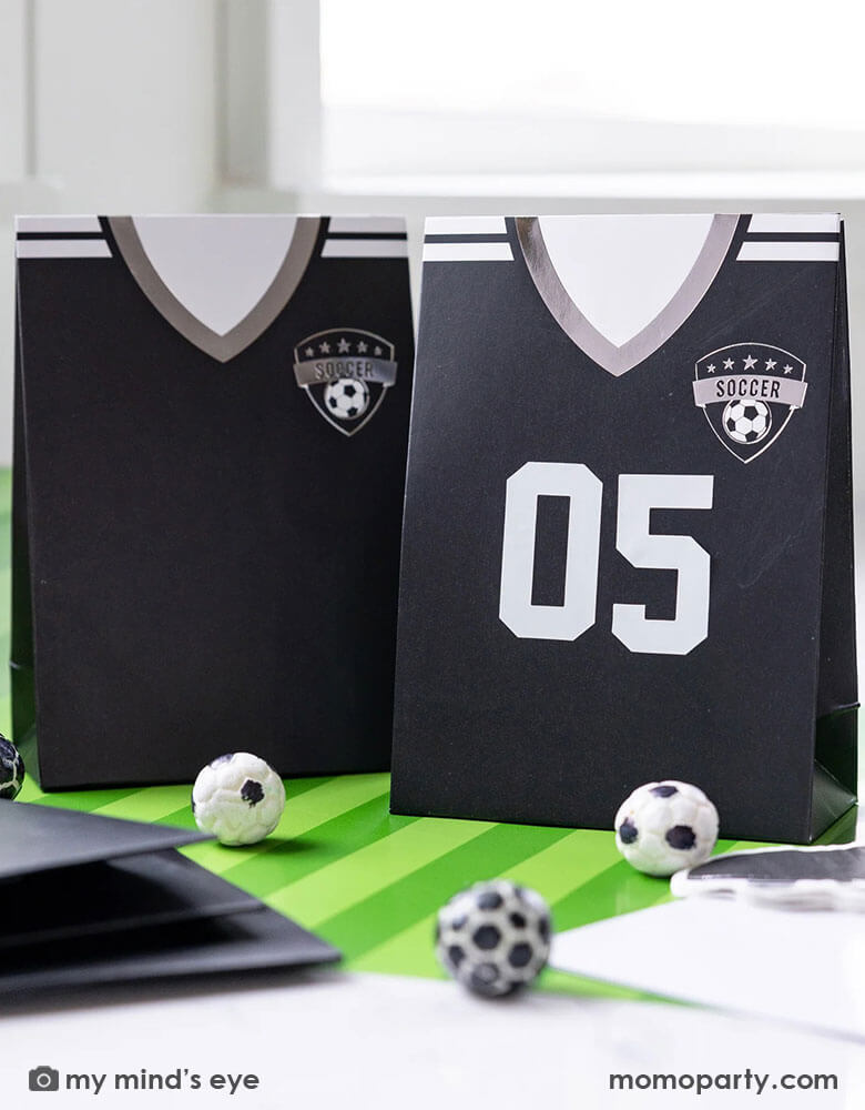 A soccer themed party table features Momo Party's black soccer jersey shaped treat bags with number stickers by My Mind's Eye. On the table there's a green striped soccer table runner and some soccer ball shaped chocolate balls, making it perfect for kid's soccer themed birthday party or championship celebration bash!