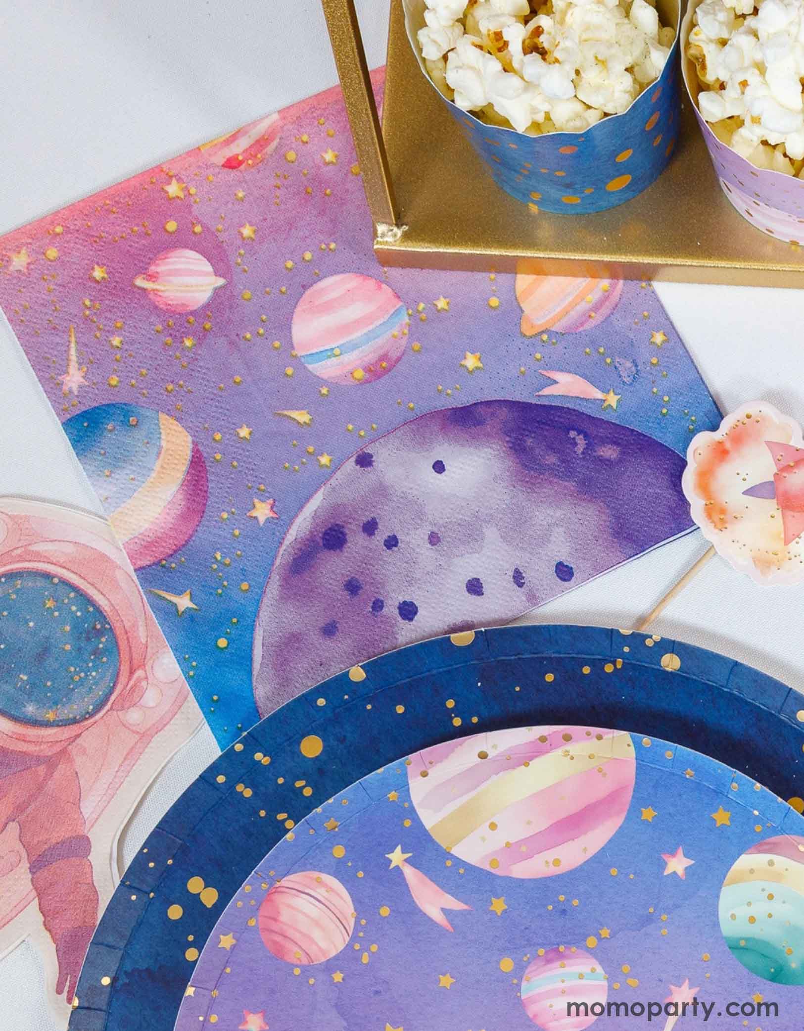 A space-themed party table with space themed tableware spread out including Momo Party's pink space napkin, the pink astronaut shaped napkin, galaxy paper plates and space themed food cups filled with popcorn on a white background.