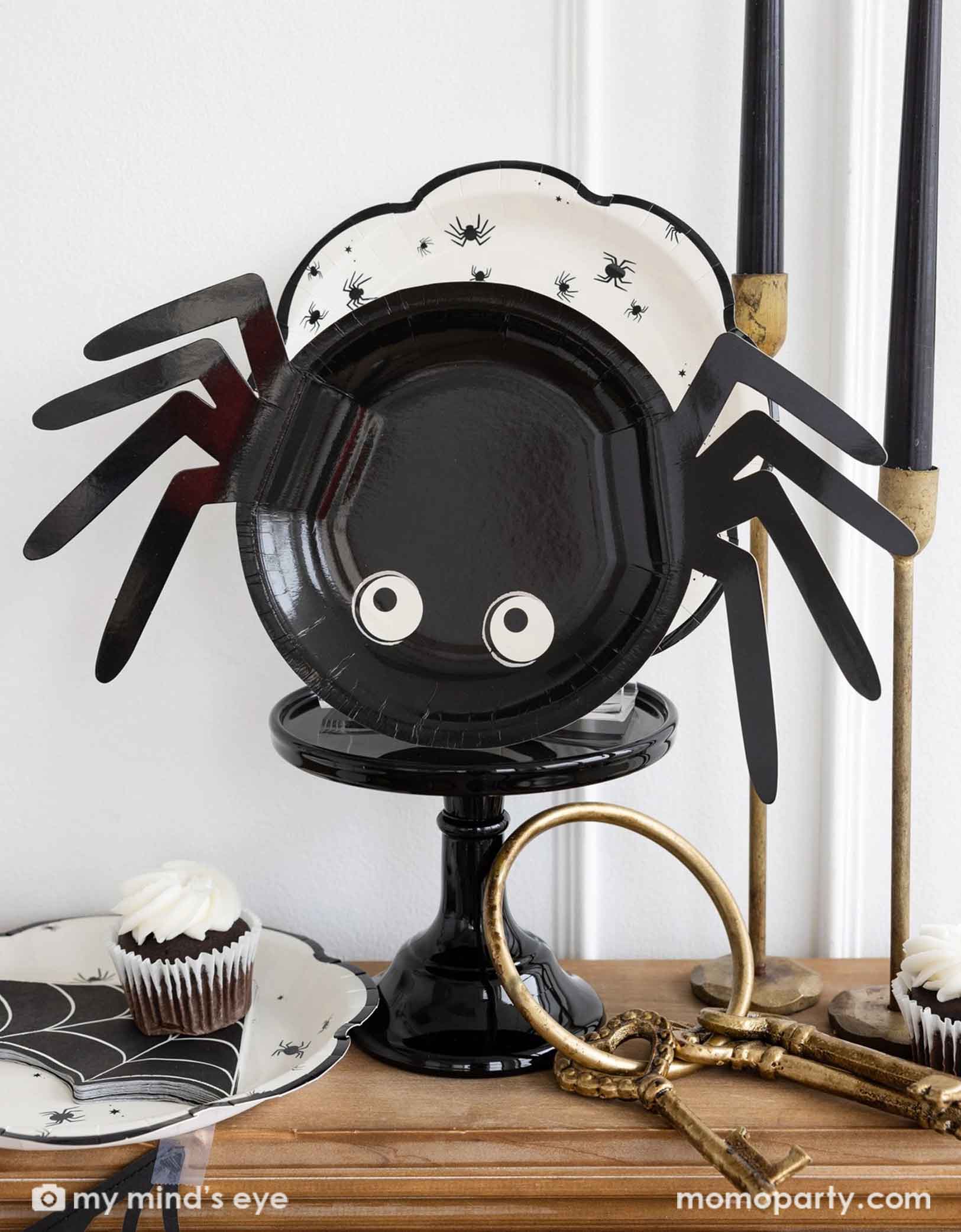 Momo Party's Spider-shaped black paper plate with white eyes, along with a large plate with spiders design on a cake stand, along surrounded by Halloween-themed decor.
