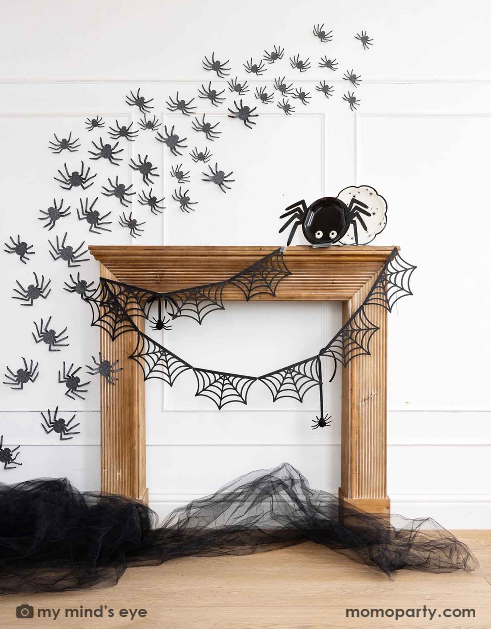 A wooden mantel against a white wall decorated with Halloween decorations from Momo Party including the spider web banner, black spider decorations on the wall and the 14" x 7" spider shaped plate and a round plate with spider designs on it. On the floor there's some black tulle on the floor, making this a kid's friendly cute Halloween decorating idea for the spooky season.