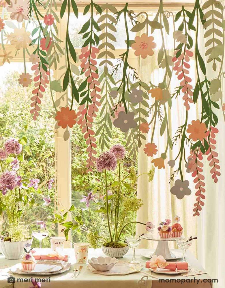 A spring inspired party table set up next to a large window with garden in the back. On the table there are some spring floral cupcakes, party crackers and party tableware, above the table hung Momo Party's floral paper backdrop which features lots of flower and foliage paper pennants, creating a wow effect to this event space.