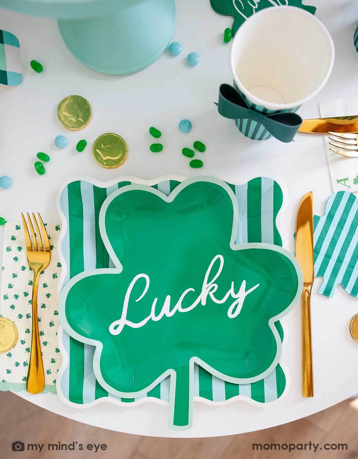 A St. Patrick's Party table setting featuring Momo Party's shamrock-shaped party plate with 'Lucky' text on it on top of a green and light blue striped square plate, with shamrock themed party napkins and party cups around on a festive table setting with gold cutlery, green candies and gold coins. A perfect inspiration for a modern St. Patrick's Day party tablescape.