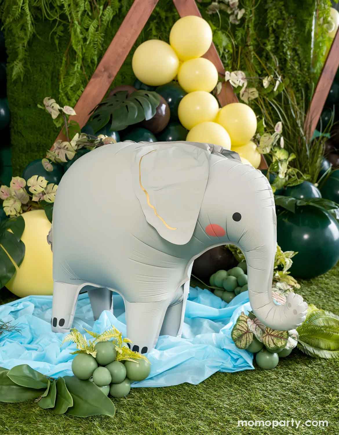 Momo Party's inflated standing elephant shaped foil party balloon displayed on a blue fabric runner with green grapes and foliage, set against a tropical green backdrop with yellow balloons at a kid's safari themed birthday party set up.