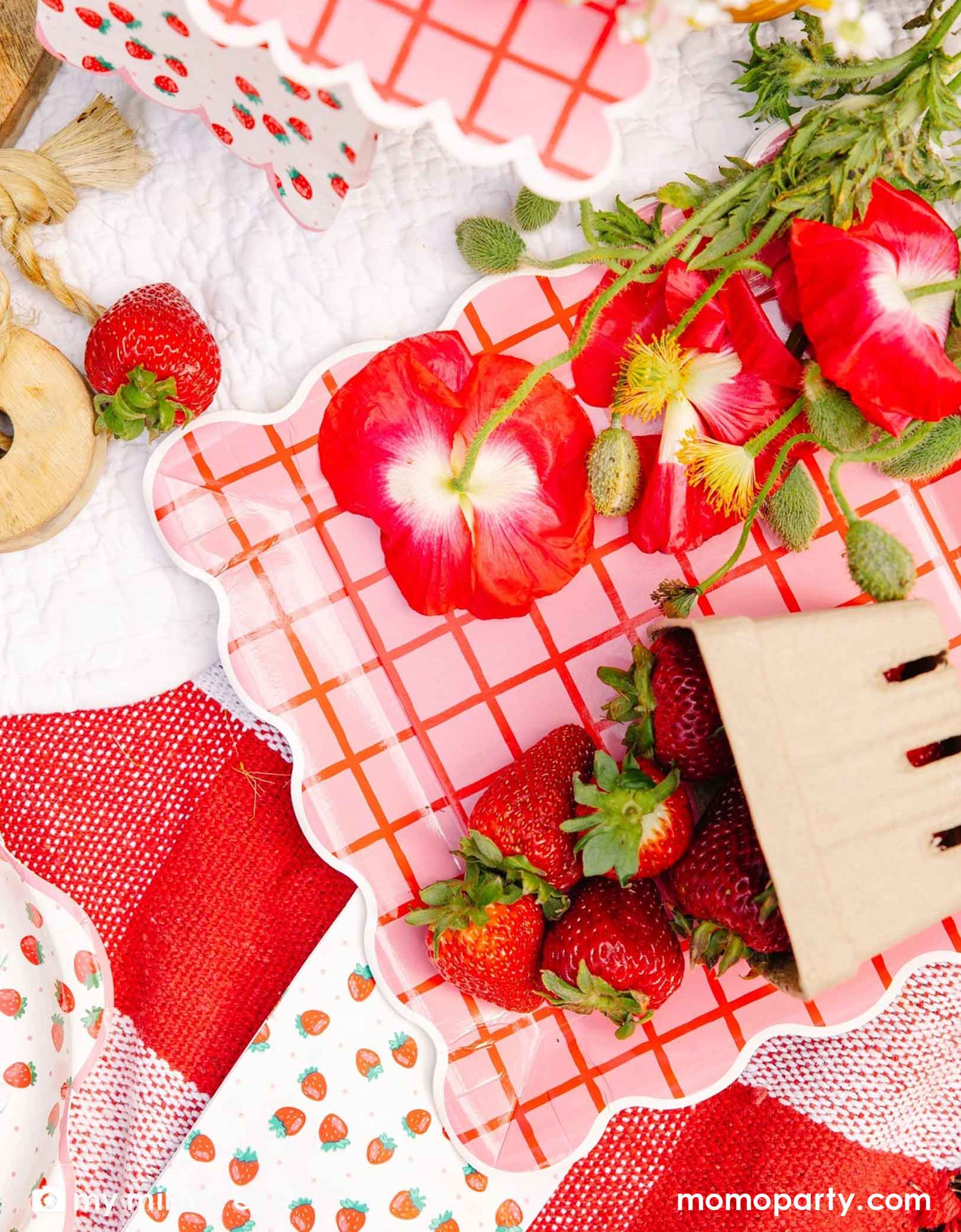 An overhead shot of a strawberry themed tablescape featuring Momo Party's strawberry tablewares including the 10" red and pink grid paper plates, strawberry patterned napkins and plates. Along with red flowers and strawberries scattered around on a white surface.