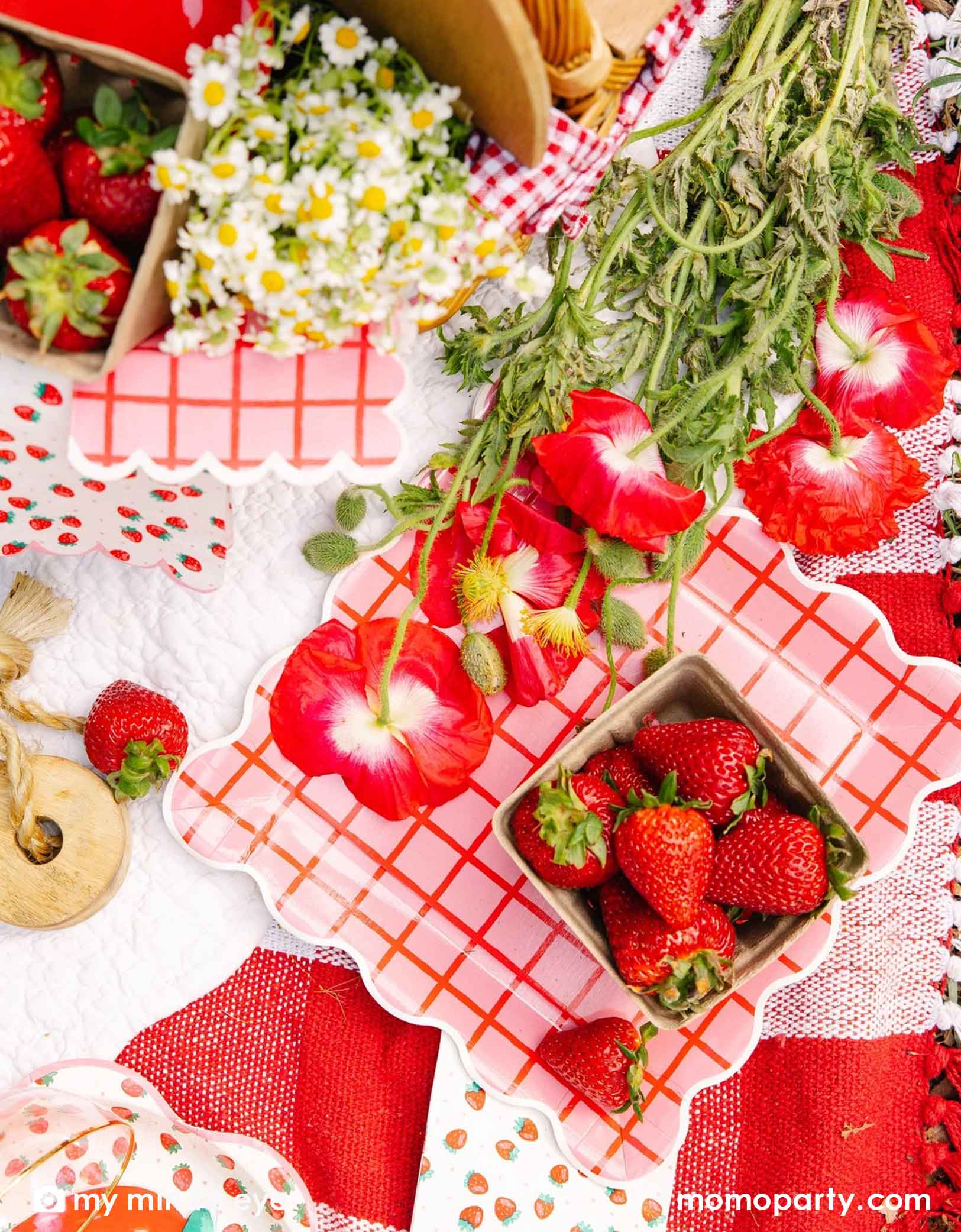An overhead shot of a strawberry themed tablescape featuring Momo Party's strawberry tablewares including the 10" red and pink grid paper plates, strawberry patterned napkins and plates. Along with red flowers and strawberries scattered around on a white surface.