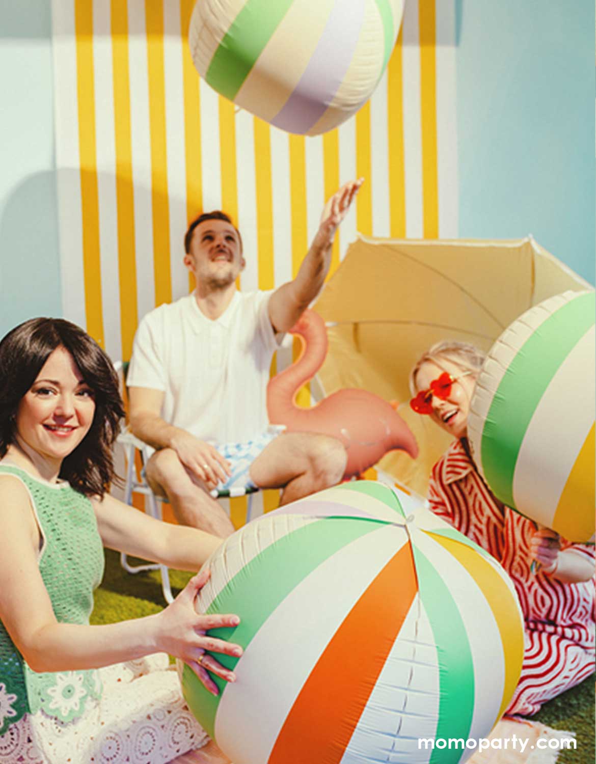 Three young people in a summer outfits in a festive summer party playing with Momo Party's 19" colorful striped beach ball shaped foil balloons against a yellow striped wall with yellow sun umbrellas around.