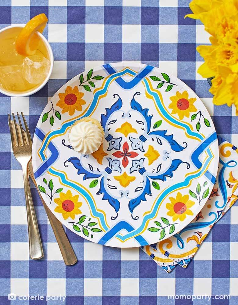 A flatlay view of a summer party table features Momo Party's navy gingham tablecover by Coterie and summer themed paper plates and napkins on it, next to the tableware there are some yellow flowers and a drink topped with an orange slice. Making this a perfect inspo for a stylish summer party tablescape.