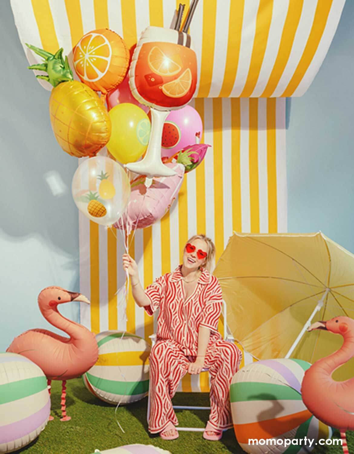 A lady in her red heart shaped sunglasses and red striped summer outfits sitting on a beach chair and holding a bunch of summer themed shaped foil balloons from Momo Party including a colorful striped beach ball balloon, a pink summer drink shaped balloon, a pineapple balloon, and multiple fruit themed balloons, along with two pink flamingo standing foil balloons and three colorful striped beach ball shaped foil balloons against a yellow striped wall with a sun umbrella next to it.