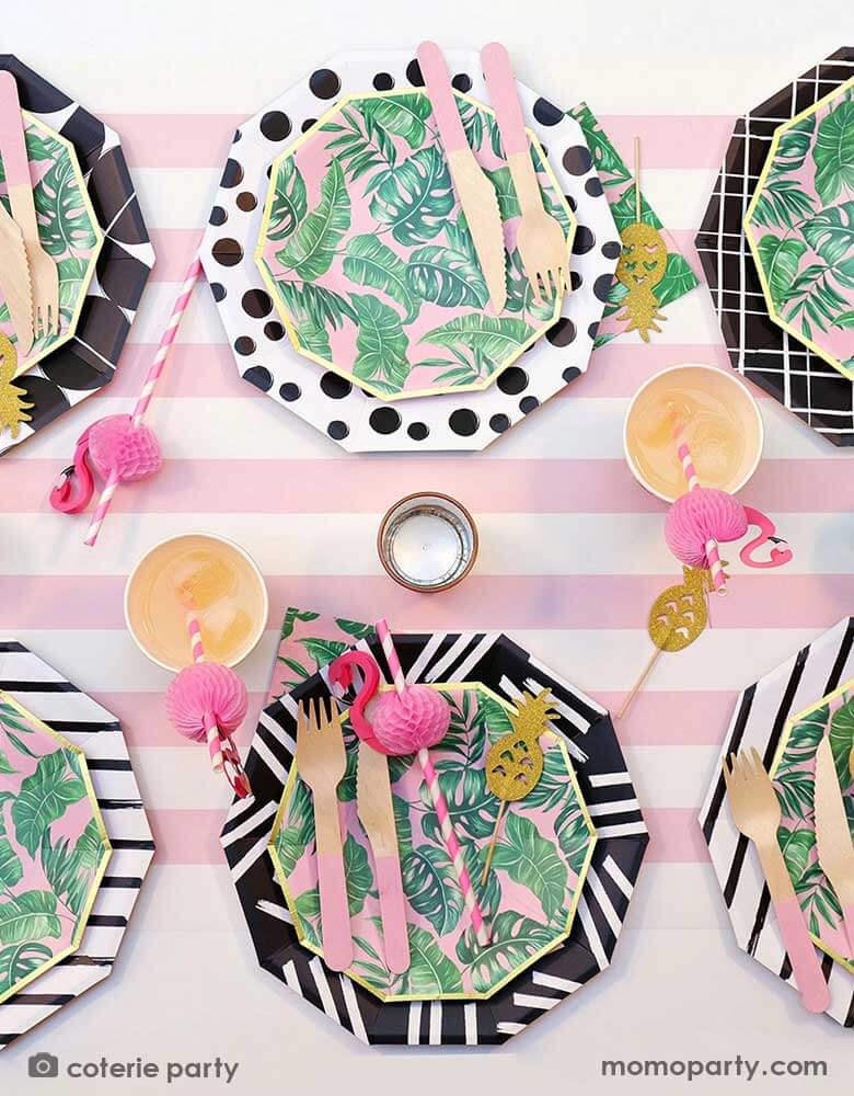 A modern summer tropical themed party tablescape features Momo Party's tropical party themed party supplies including palm leaf plates, napkins, pink wooden utensil set and flamingo honeycomb paper straws in the party cups. All these are on a pink striped table runner, making this a festive yet modern look for a summer party!