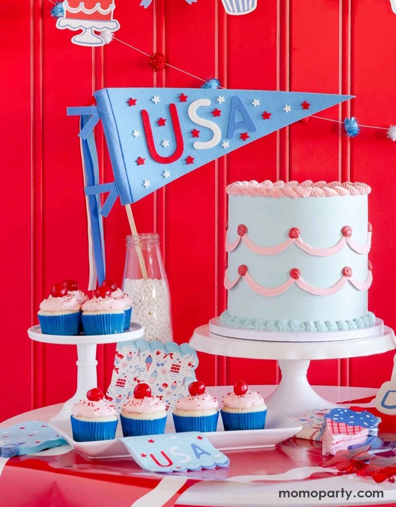 A festive patriotic party set up featuring various Fourth of July themed party decors from Momo Party including party banner with summer themed party pennants including popsicles, ice cream, cupcake, cakes, and star shaped sunglasses in red, blue and white colors. On the table there's a vintage pastel blue buttercream cake along with some cupcakes with cherries on top, along with a USA felt pennant and patriotic party tableware including party plates, napkins and a table runner, for a fun summer party.