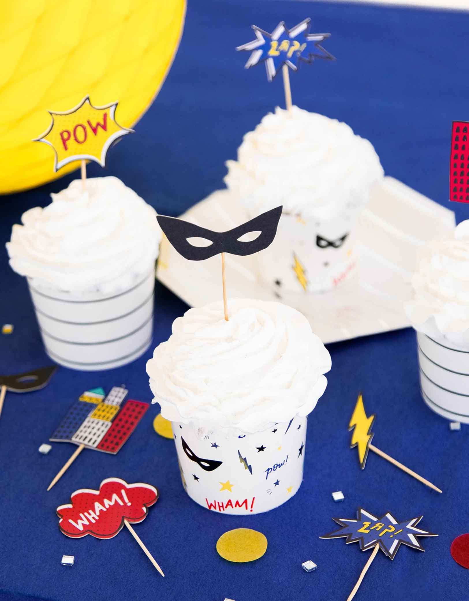 A blue party table features cupcakes decorated with Momo Party's superhero themed cupcake kit featuring superhero masks, city buildings, lightning bolt, and action words on sticks .