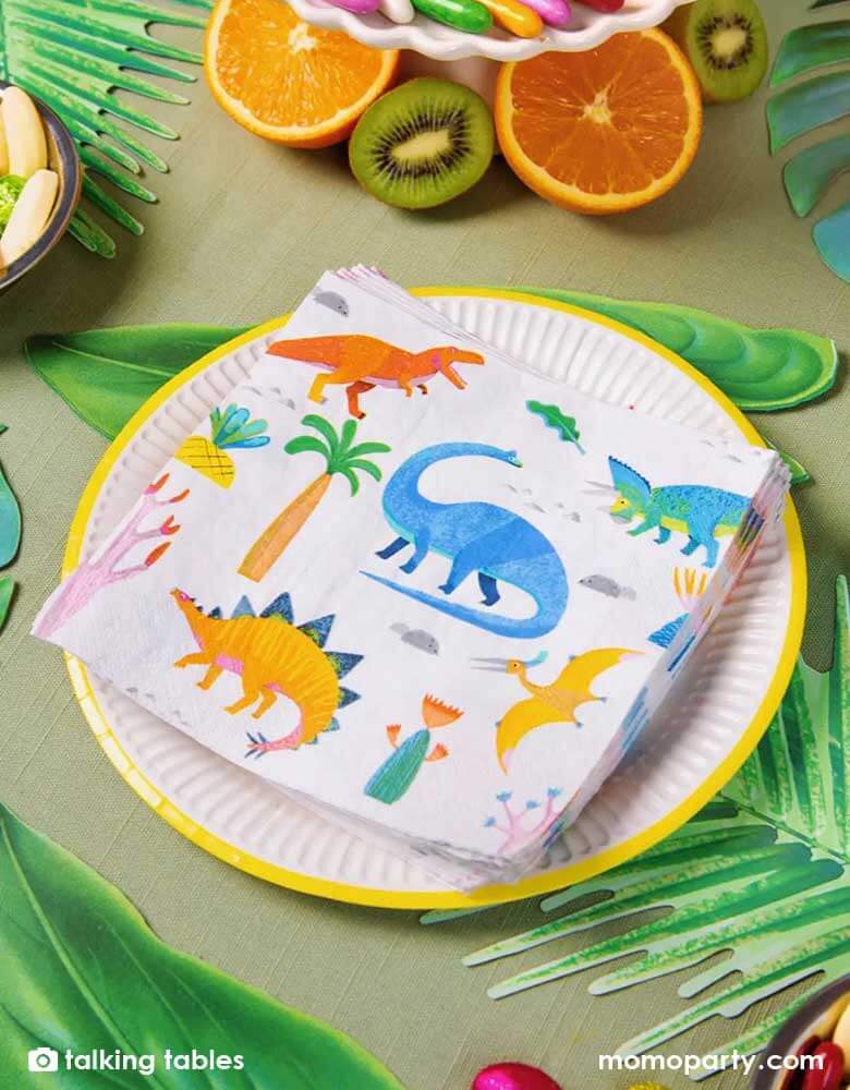 A prehistorical jungle dinosaur themed party table decorated with palm tree leaves and tropical fruits, this table features Momo Party's Momo Party's 6.5 x 6.5 inches dinosaur party napkins by Talking Tables. Comes in a set of 20 napkins, featuring 5 different jurassic dinosaurs, these colorful napkins are perfect for catching crumbs or mopping up drink spills. A practical addition to a kids dinosaur birthday party.
