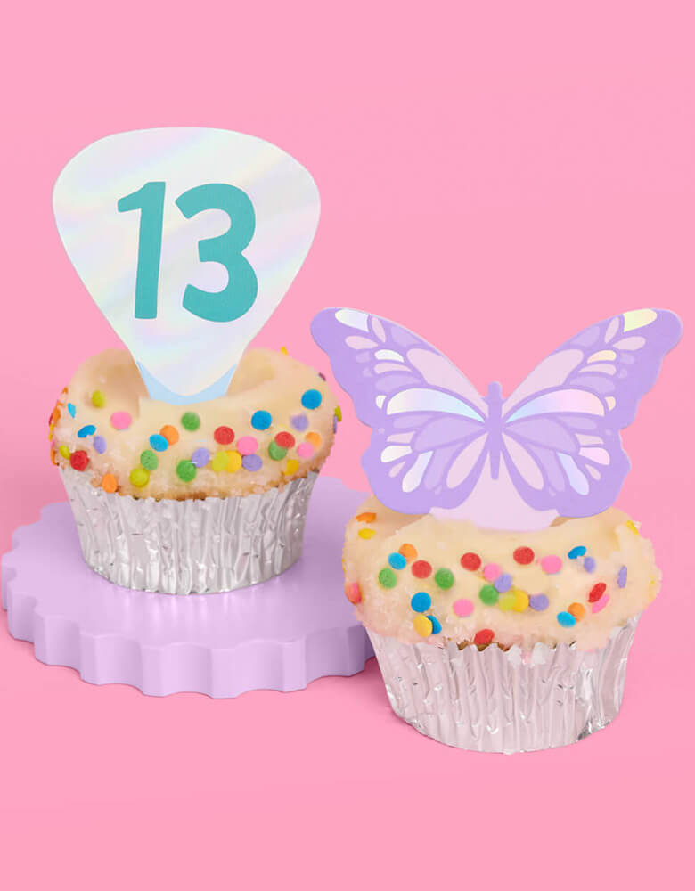 Two birthday cupcakes adorned with Momo Party's Era's Party Cupcake Toppers by Xo, Fetti. Comes in a set of 24, these Taylor Swift-themed dessert/cupcake toppers are with iconic designs including butterflies, red lips, disco balls, cassettes, guitar picks and heart hand symbols. They're perfect for to celebrate your Swiftie girl's special birthday.