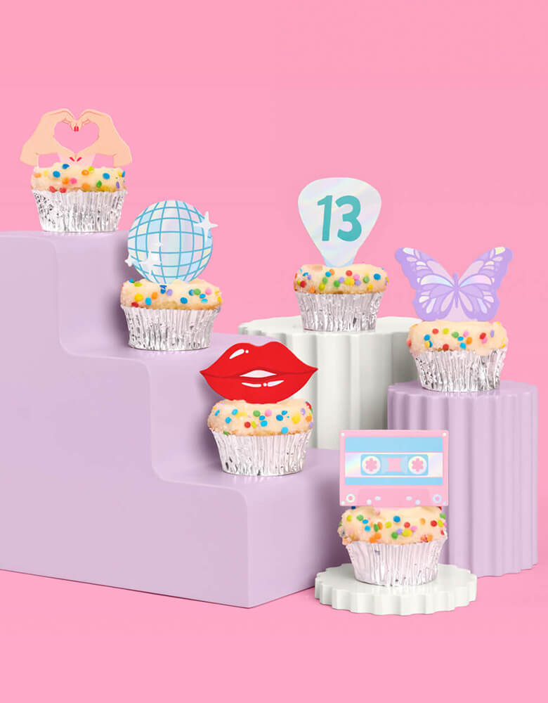 A few birthday cupcakes adorned with Momo Party's Era's Party Cupcake Toppers by Xo, Fetti on a modern lilac cupcake stand. Comes in a set of 24, these Taylor Swift-themed dessert/cupcake toppers are with iconic designs including butterflies, red lips, disco balls, cassettes, guitar picks and heart hand symbols. They're perfect for to celebrate your Swiftie girl's special birthday.