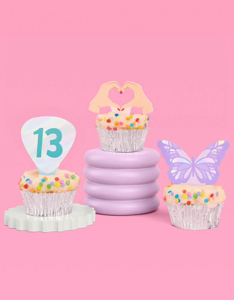 Three birthday cupcakes adorned with Momo Party's Era's Party Cupcake Toppers by Xo, Fetti. Comes in a set of 24, these Taylor Swift-themed dessert/cupcake toppers are with iconic designs including butterflies, red lips, disco balls, cassettes, guitar picks and heart hand symbols. They're perfect for to celebrate your Swiftie girl's special birthday.