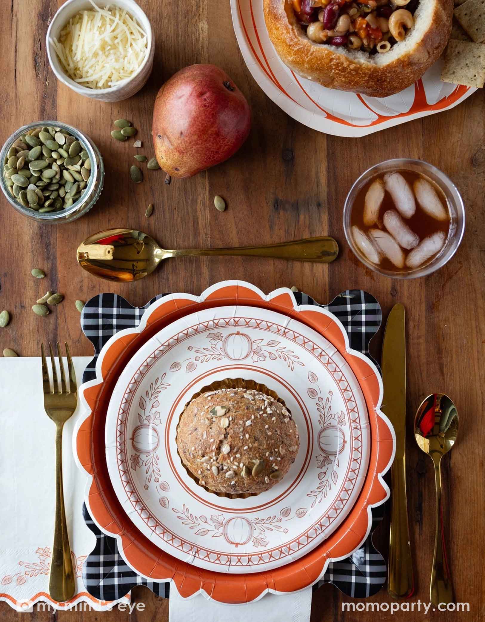 A festive Thanksgiving friendsgiving themed rustic table setting with pumpkin themed plates, bowls, napkins drinks, and gold utensils from Momo Party on a wooden board. With decorative fall themed items including pumpkins, pumpkin seeds, cheese, fall floral and fruit branches, it creates a perfect inspo for a Thanksgiving party, friendsgiving or fall, harvest themed gathering in autumn.