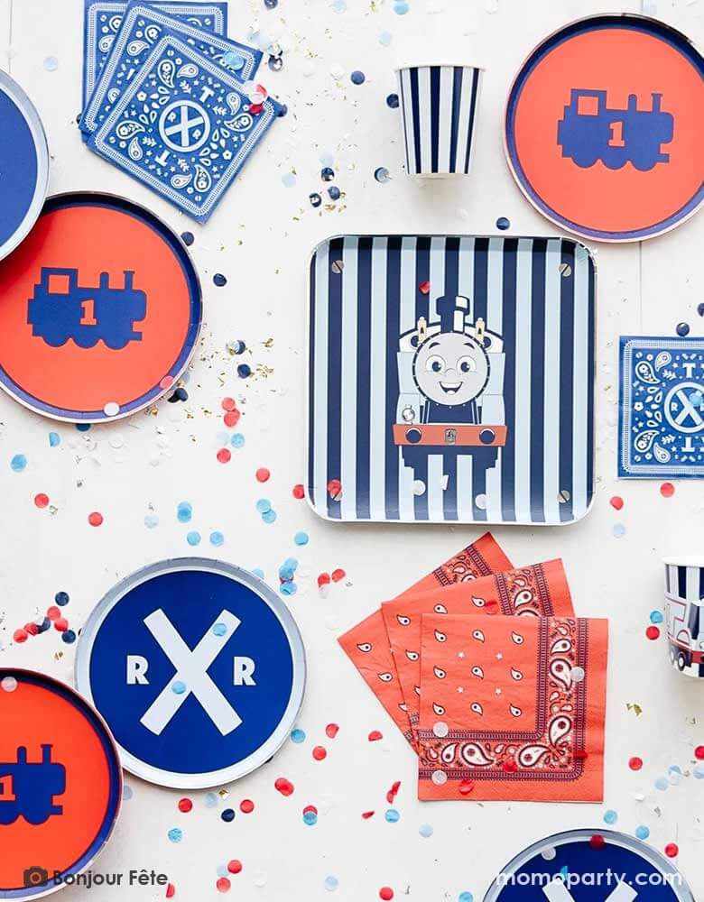 A table full of Thomas and Friend's themed party supplies from Momo Party by Bonjour Fete including blue striped square dinner plate with Thomas illustration on it, assorted side plates in blue and red, bandana inspired paper napkins in red and blue and Thomas 3D blue striped party cups, along with blue and red confetti spread out this is a perfect inspo for a modern kid's train themed Thomas and Friends birthday party!