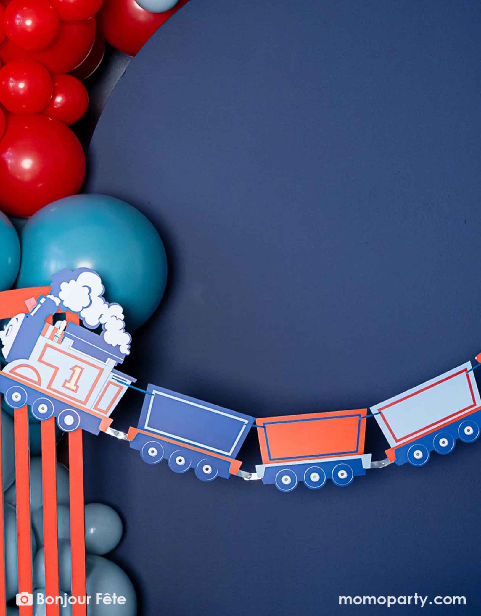 A close-up shot of Momo Party's Thomas the Train and Friends party garland hung on a blue wall which is decorated with a balloon garland in the classic train colors of blue and red.
