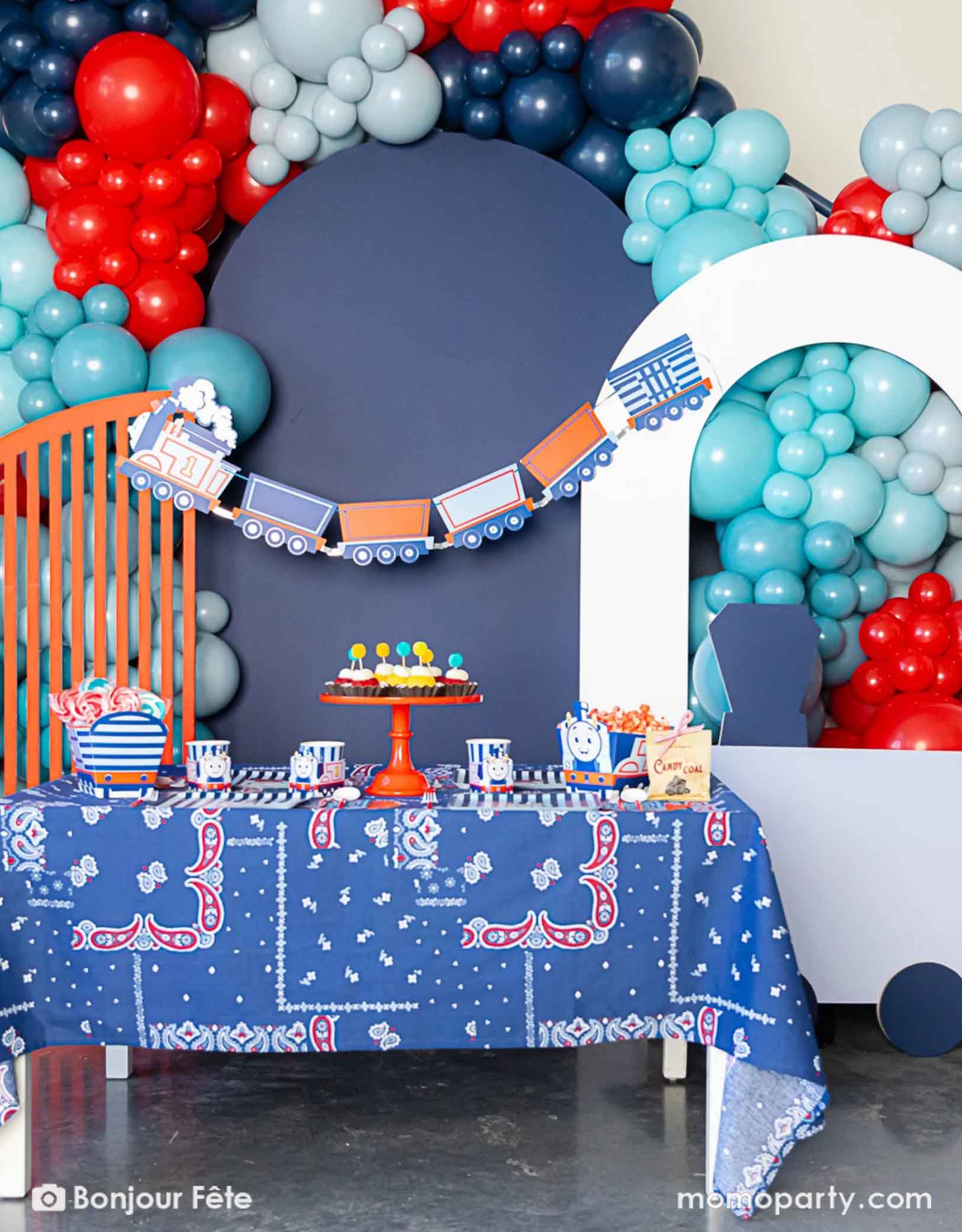 An adorable and festive Thomas the Train and Friends themed train party set up featuring Momo Party's Thomas the Train themed party supplies including party tableware of plates, cups, napkins treat boxes and party garland. The party table is covered with bandana inspired tablecloth to match the theme. In the middle of the table there's a cake stand holding cupcakes in yellow and red colors topped with pom pom toppers. In the back of is a blue & red balloon garland, a perfect inpo for kid's train party.