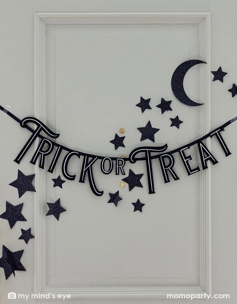 A gray front door decorated with Momo Party's 4ft trick or treat felt banner by My Mind's Eye. Around the felt banner are some black glittered stars and moon wall paper decorations, making this a great inspo for Halloween home decoration this spooky season!