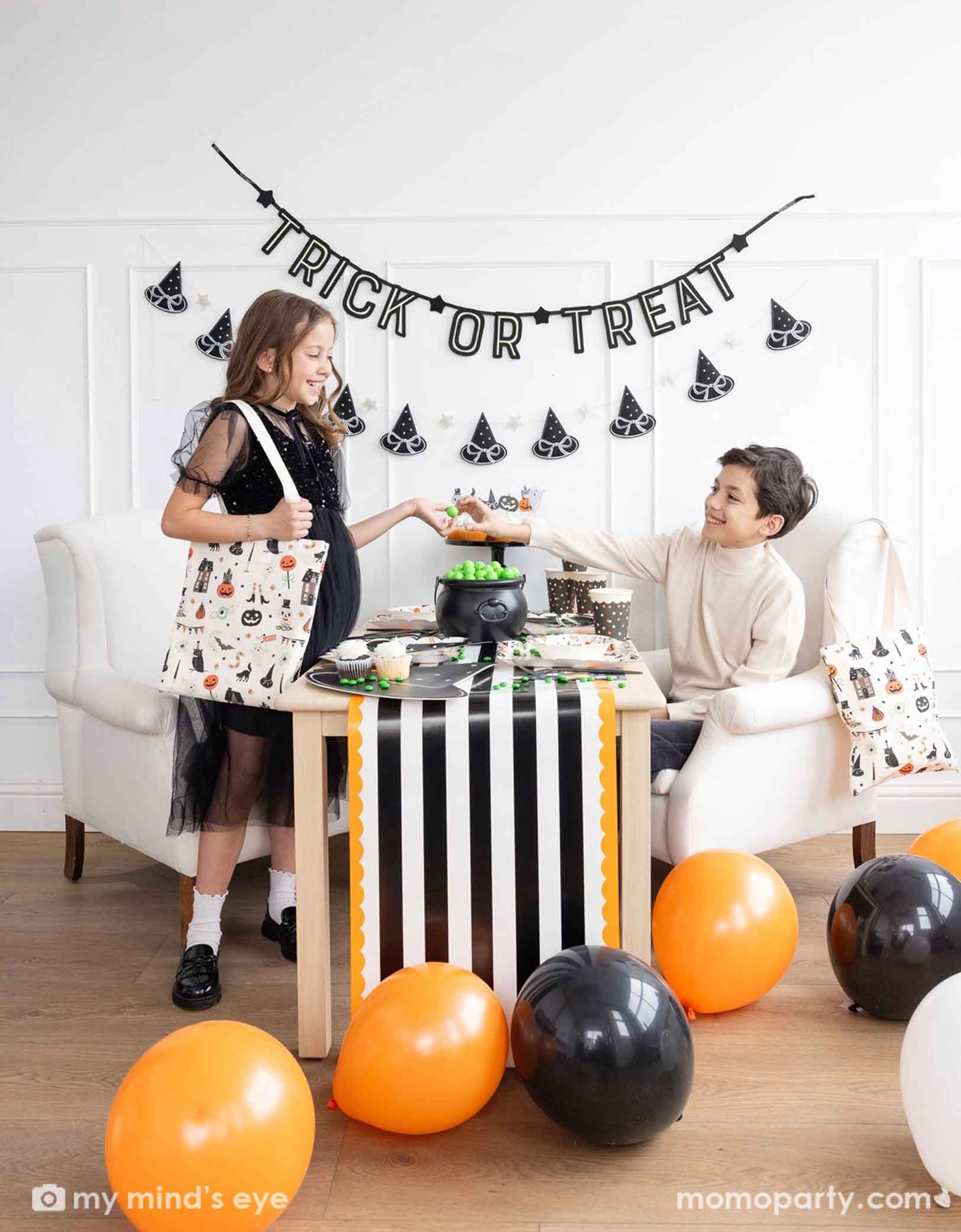 Two children at a Halloween-themed party with Momo Party's 'Trick or Treat' banner and the witch's hat garland hung on the white wall. There's a table decorated with multiple Halloween party supplies by Momo Party including witch's hat shaped bamboo tray, party cups and orange, black and white striped table runner draping from the table which some orange and black balloons next to. A cute and simple set up for kids to celebration Halloween at home.