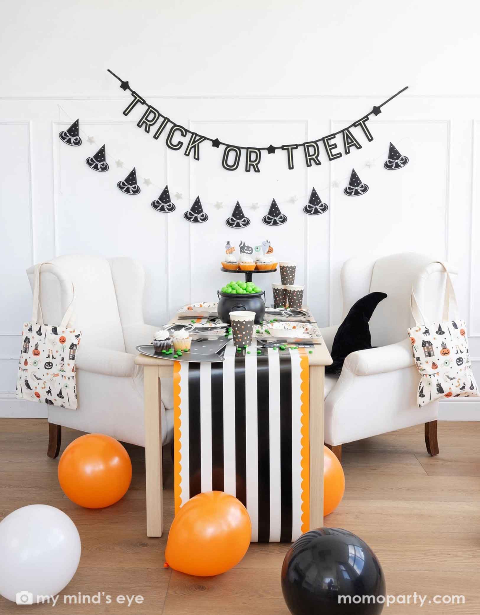 A Halloween-themed party with Momo Party's 6 ft 'Trick or Treat' glow in the dark banner and the witch's hat garland hung on the white wall. There's a table decorated with multiple Halloween party supplies by Momo Party including witch's hat shaped bamboo tray, party cups and orange, black and white striped table runner draping from the table which some orange and black balloons next to. A cute and simple set up for kids to celebration Halloween at home.