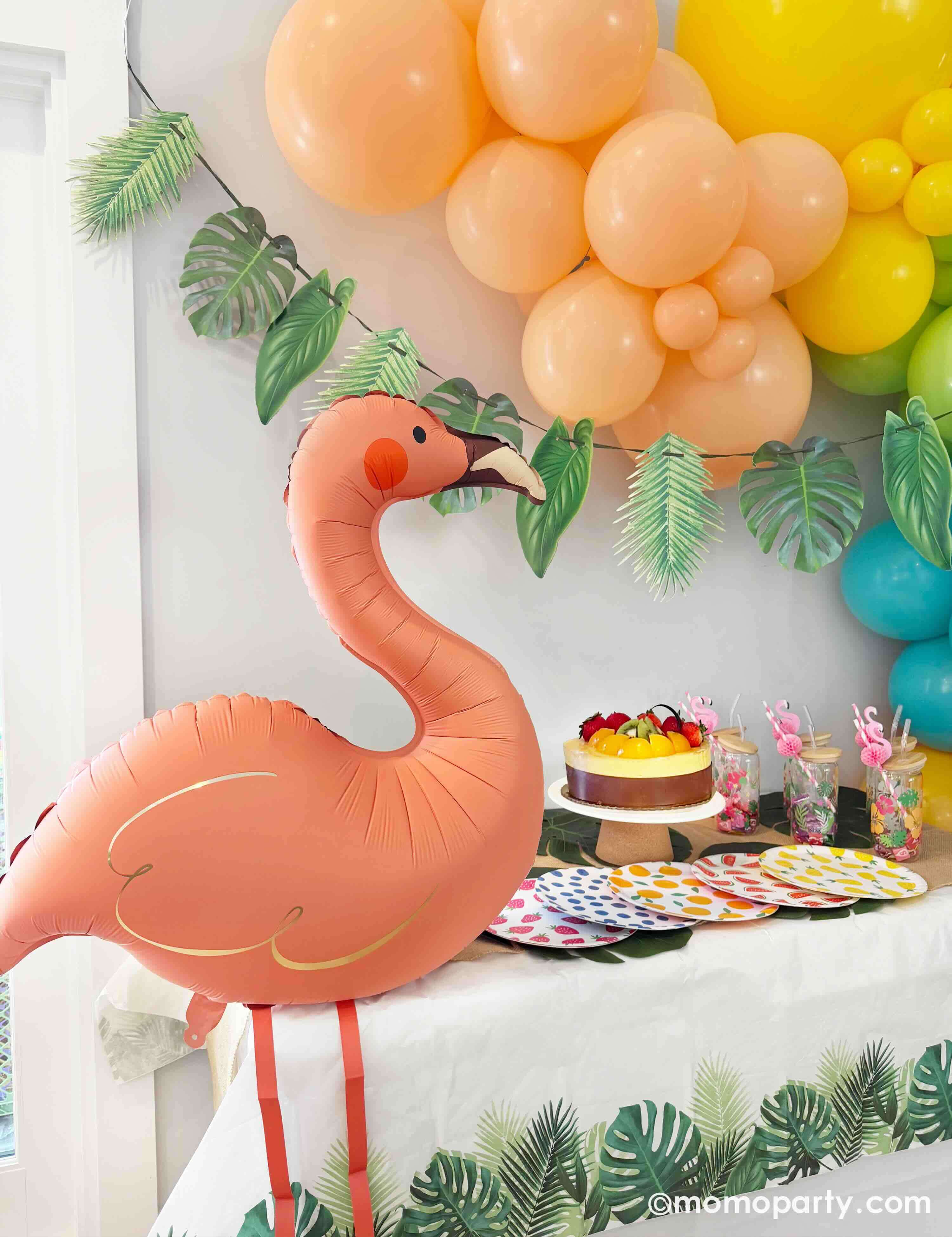 A tropical/island/aloha themed birthday party decorative setup by Momo Party with a festive balloon garland in tropical inspired colors of peach, yellow, lime green, Caribbean blue, and orange, adorned with a palm leaf party garland, along with a flamingo shaped foil balloon, and tropical-themed fruit and palm tree tableware from Momo Party.