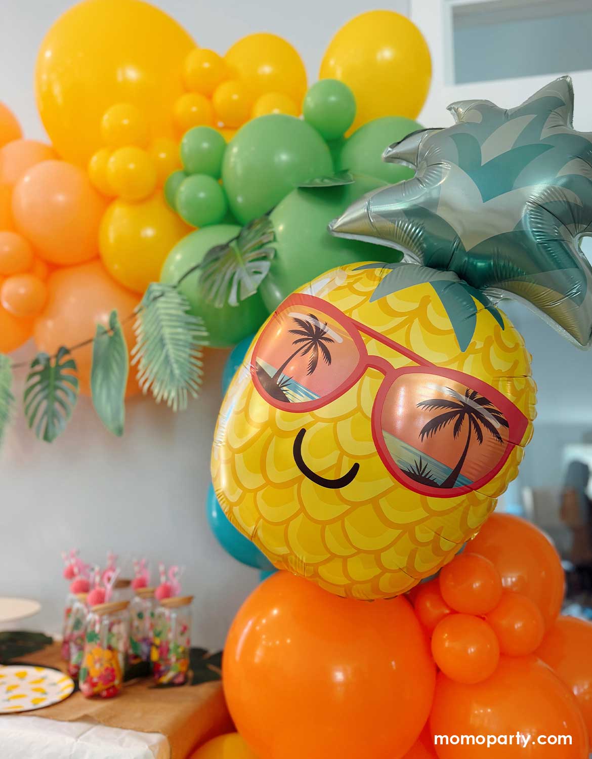 A tropical/island/aloha themed birthday party decorative setup by Momo Party with a festive balloon garland in tropical inspired colors of peach, yellow, lime green, Caribbean blue, and orange, adorned with a palm leaf party garland, along with a pineapple shaped foil balloon with sunglasses, and tropical-themed fruit and palm tree tableware from Momo Party on the dessert table. A fun and festive set up for kids summer themed parties.