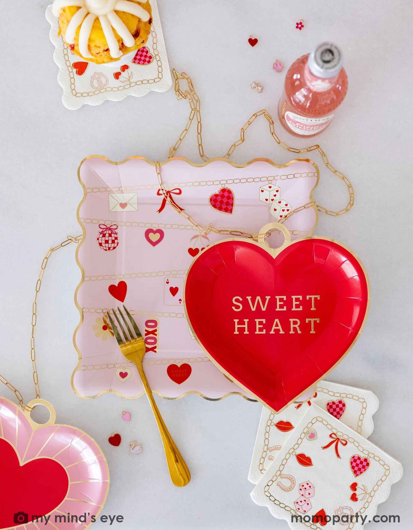 An overhead shot of a Valentine's Day party tablescape featuring various Valentine's Day party supplies from Momo Party including the red heart shaped plate with 'Sweet Heart' text and Valentines Charm Scalloped Square Plate and Valentines Charm Small Napkins by My Mind's Eye, surrounded by decorative Valentine's items, charms, snacks and drinks on a light background.