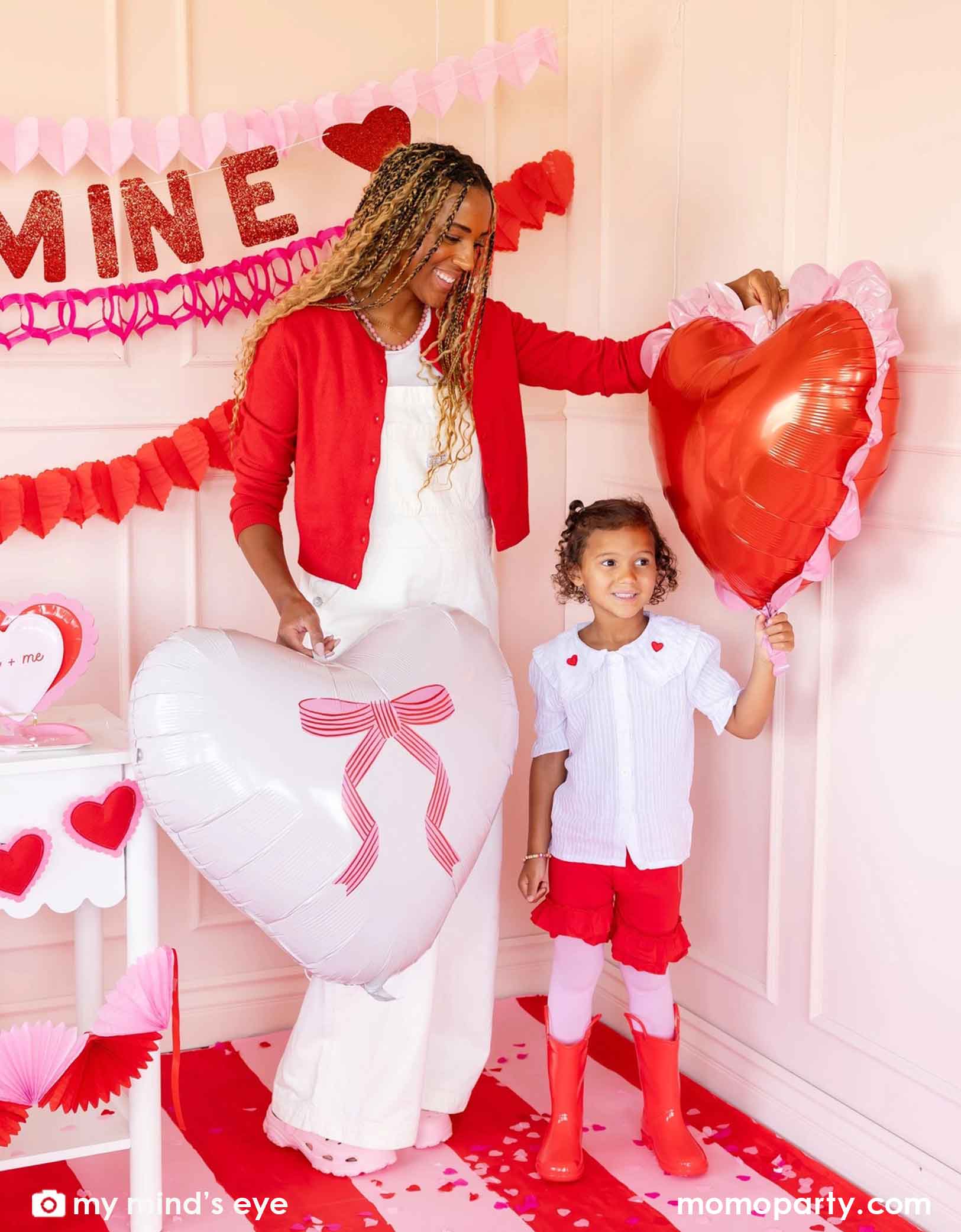 A mom and her child in red and pink outfits holding Momo Party's 22" heart-shaped balloons by My Mind's Eye. With 'BE MINE' party banner and red and pink tissue paper garlands in the back on a pink wall. In the front there's a dessert table filled with Valentine's Day party supplies and heart shaped decorations. A perfect inspiration for a sweet Valentine's Day celebration with family and kids.