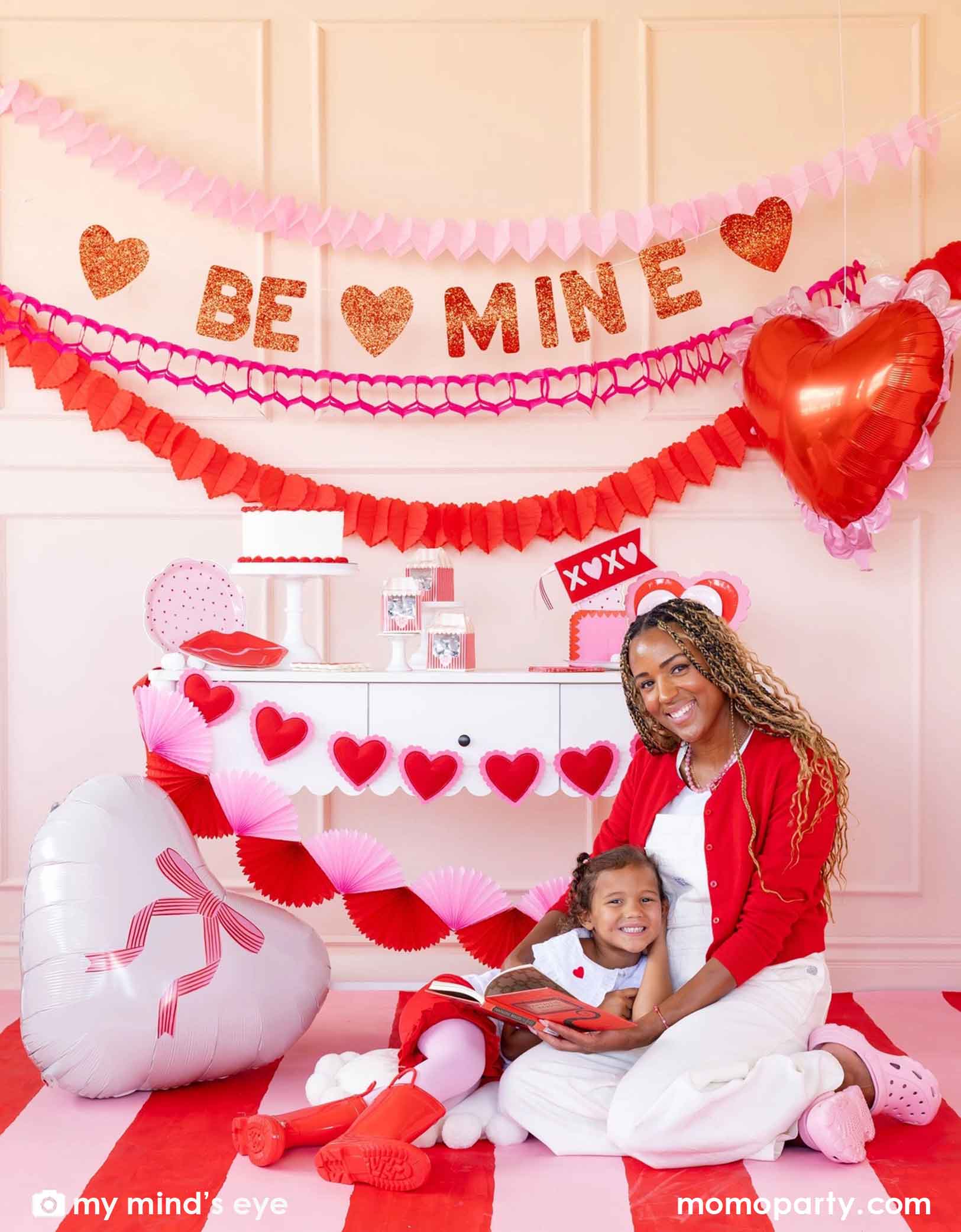 A mom and her kid sitting on a red and white striped rug at a Valentine's Day party set up featuring various Momo Party's Valentine's Day party supplies and decorations including 22" heart-shaped balloons by My Mind's Eye, 'BE MINE' party banner, red and pink tissue paper garlands in the back on a pink wall. In the front there's a dessert table filled with Valentine's Day party supplies and heart shaped banner decorations. A perfect inspiration for a sweet Valentine's Day celebration with family and kids.