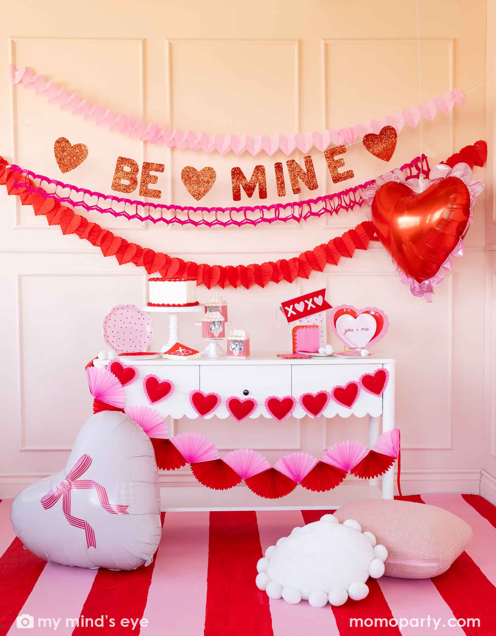 A festive Valentine's Day party set up featuring various Momo Party's Valentine's Day party supplies and decorations including two 22" heart-shaped balloons by My Mind's Eye, 'BE MINE' party banner, red and pink tissue paper garlands in the back on a pink wall. In the front there's a dessert table filled with Valentine's Day party supplies and heart shaped banner decorations. A perfect inspiration for a sweet Valentine's Day celebration with family and kids.