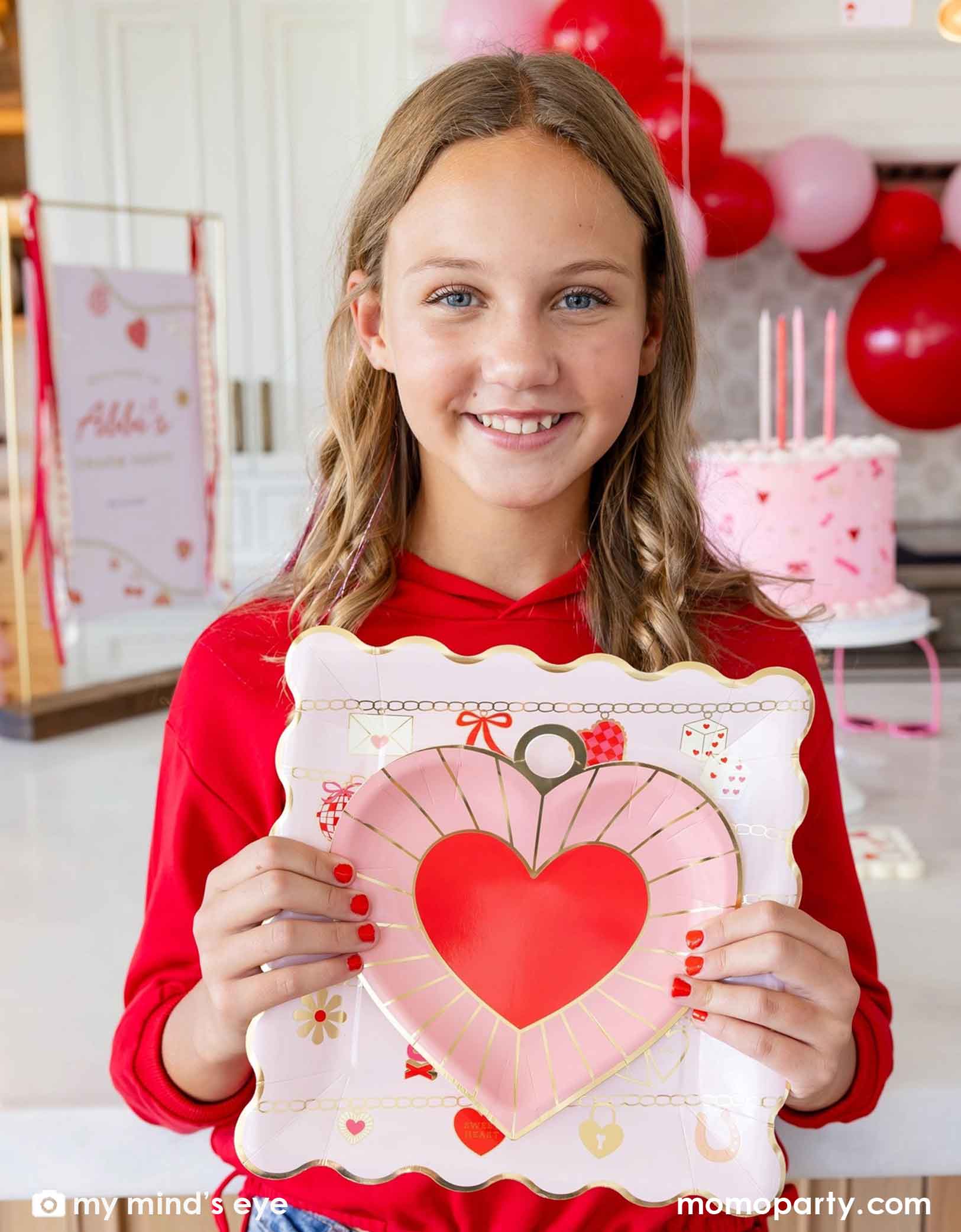 A young blonde girl in a Valentine's Day party scene holding Momo Party's pink heart-shaped plate and pink square plate with Valentine's Day balloon garland and a pink buttercream cake in the background.