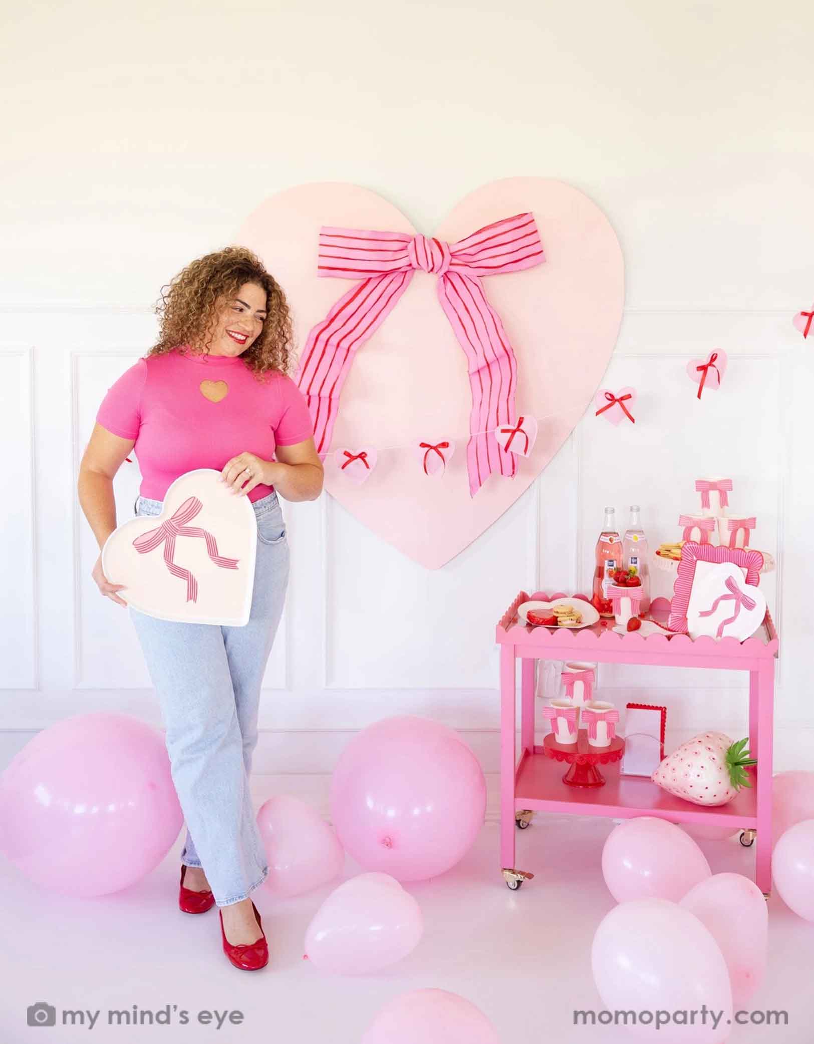 A lady with a hot pink top with red lipstick and red high heels holding Momo Party's 15" x 15" Bow Heart Tray by My Mind's Eye. She is standing next to a pink cart filled with Valentine's Day party supplies including plates, cups, napkins, along with drinks and snacks. In her back there's a giant pink heart shaped decoration with ribbon bow on the wall. With a floor of pink balloons, this makes a great scene for a sweet Valentine's Day celebration.
