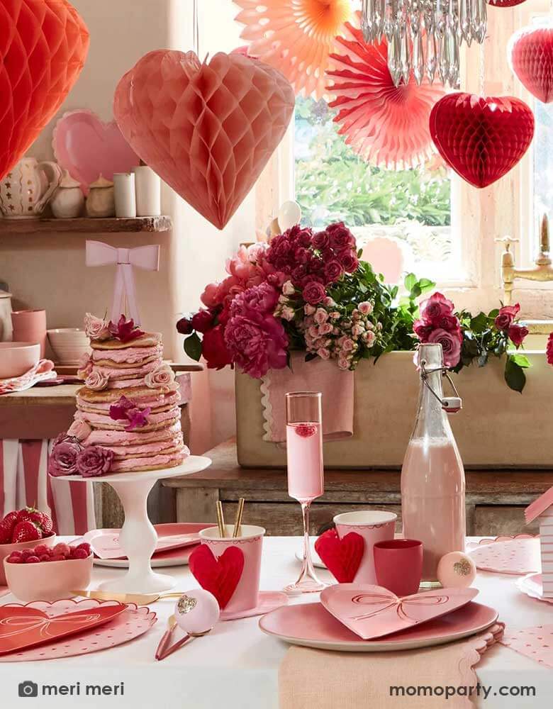 A festive Valentine's Day party featuring a table filled with Momo Party's red and pink heart shaped party supplies including the scallop edged dinner plates, napkins, heart with bow plates and heart honeycomb party cups, in the middle is a pink pancake tower adorned with pink and red flowers topped with a pink bow. Above the table, there are heart shaped honeycombs and paper fans hanging from the ceiling next to a big window, making this a great inspo for Valentine's Day celebration.