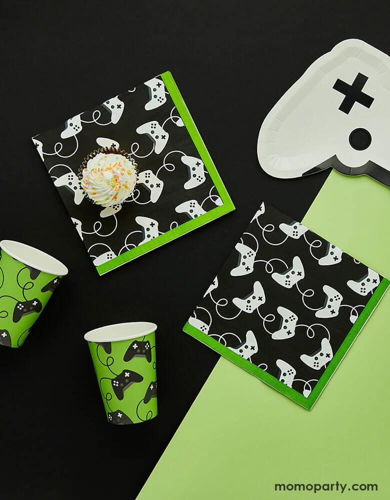 A video game themed party collection from Momo Party by Hooty Balloo including two green video game controller motif party cups, and two black game controller motif large napkins with green edge with a cupcake on them, and a white video game controller shaped paper plate, all on a classic black and green color blocked background, perfect for any game events or a kid's video game themed birthday party.