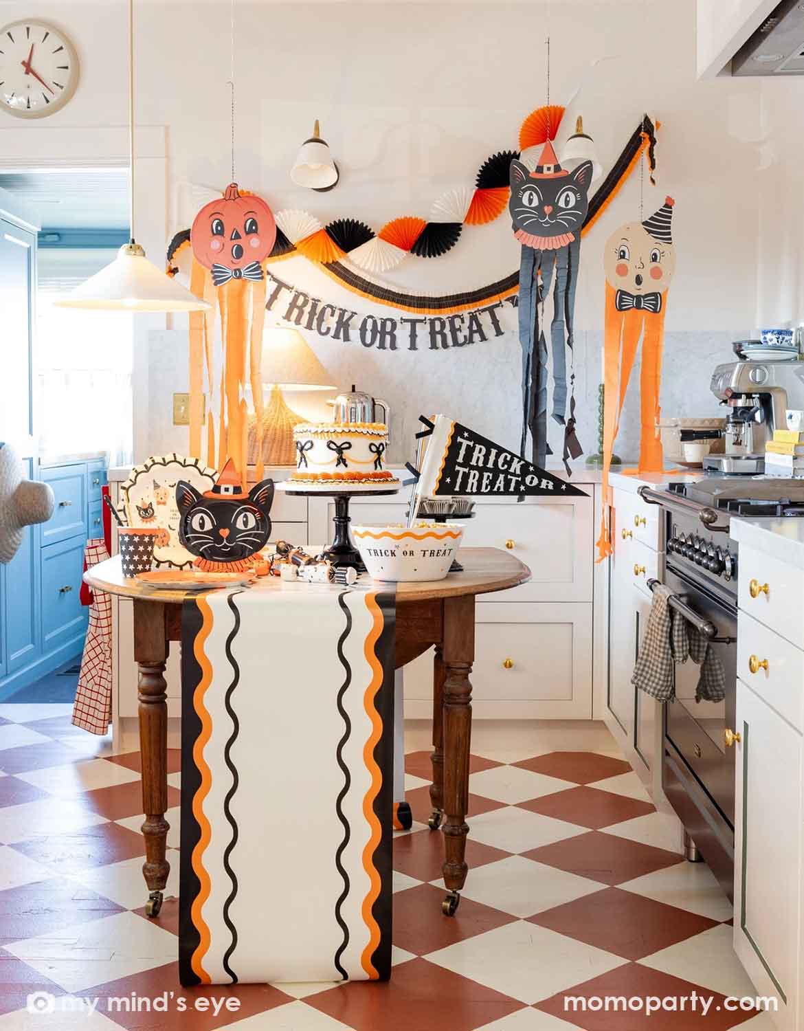 A modern kitchen with vintage accents decorated for Halloween featuring various vintage Halloween themed party supplies and decorations from Momo Party including the huge Halloween character hanging decorations, trick or treat banner, bunting garlands, and a table with a vintage Halloween-themed cake, a bowl of candy corn, and vintage Halloween themed party plates, serving bowls, cups, napkins, cupcake toppers and a table runner, making this a fun and festive set up for kids and family in the spooky season.