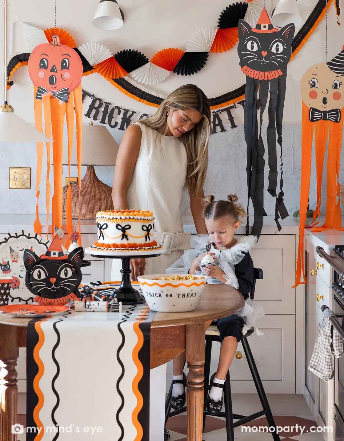 A woman and a young girl celebrate Halloween in a decorated kitchen featuring various vintage Halloween themed party supplies and decorations from Momo Party including huge hanging decorations, trick or treat banner, bunting garlands, and a table with a Halloween-themed cake, a bowl of candy corn, and vintage Halloween themed party plates, serving bowls, cups, napkins, cupcake toppers and a table runner, making this a fun and festive set up for kids and family in the spooky season.