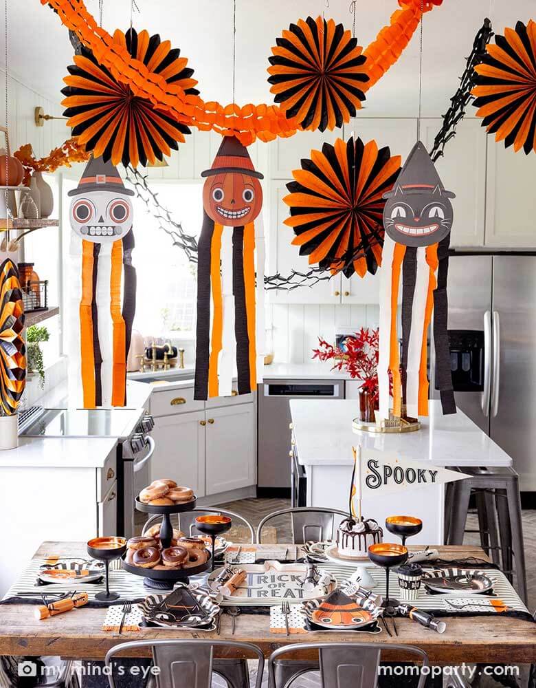 A modern kitchen that's decorated with vintage inspired Halloween party supplies form Momo Party including the Halloween hanging characters of a black cat, a pumpkin and a skull, which around them hung the oversized orange and black paper fans and tissue decorations. On the table there are matching tableware including party plates, napkins, cups and party crackers in the vintage style. With the "Spooky" felt party pennant, cakes and donuts, this creates a festive vibe for a fun Halloween celebration.
