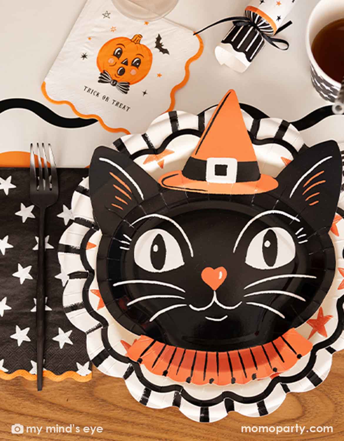 Halloween party table setting featuring various Halloween party supplies from Momo Party including a black cat face shaped paper plate with an orange witch hat and collar, a black napkin with white stars, a white bib with an orange pumpkin and the words 'TRICK OR TREAT', and a cup of coffee on a wooden table.