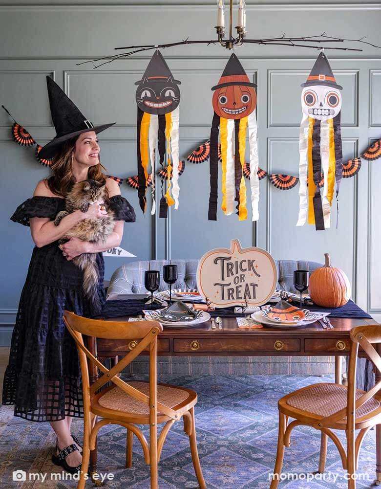 A lady dressed as a witch holding her cat in a Halloween party set up featuring vintage Halloween party supplies and decorations from Momo Party including the giant vintage Halloween character hanging decoration streamers and the black and orange bunting banner on the wall as the backdrop. On the table, there's a huge pumpkin shaped bamboo serving plate that says trick or treat on it. With vintage Halloween themed plates, napkins and glasses. This makes a great inspo for a fun Halloween gathering at home.