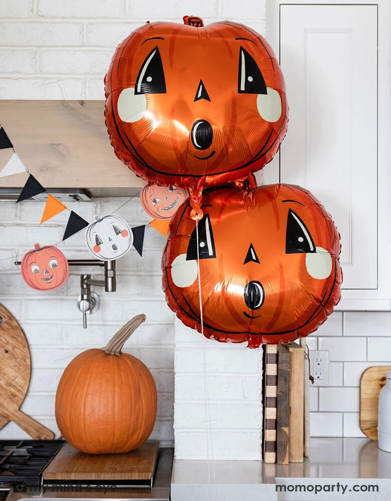 A modern white kitchen decorated for Halloween with Momo Party's vintage Halloween party supplies by My Mind's Eye including pumpkin shaped foil balloons, black, white and orange pennant flags and and paper banner. With a large pumpkin next to the stove, this makes a festive Home decoration to celebrate the spooky season.