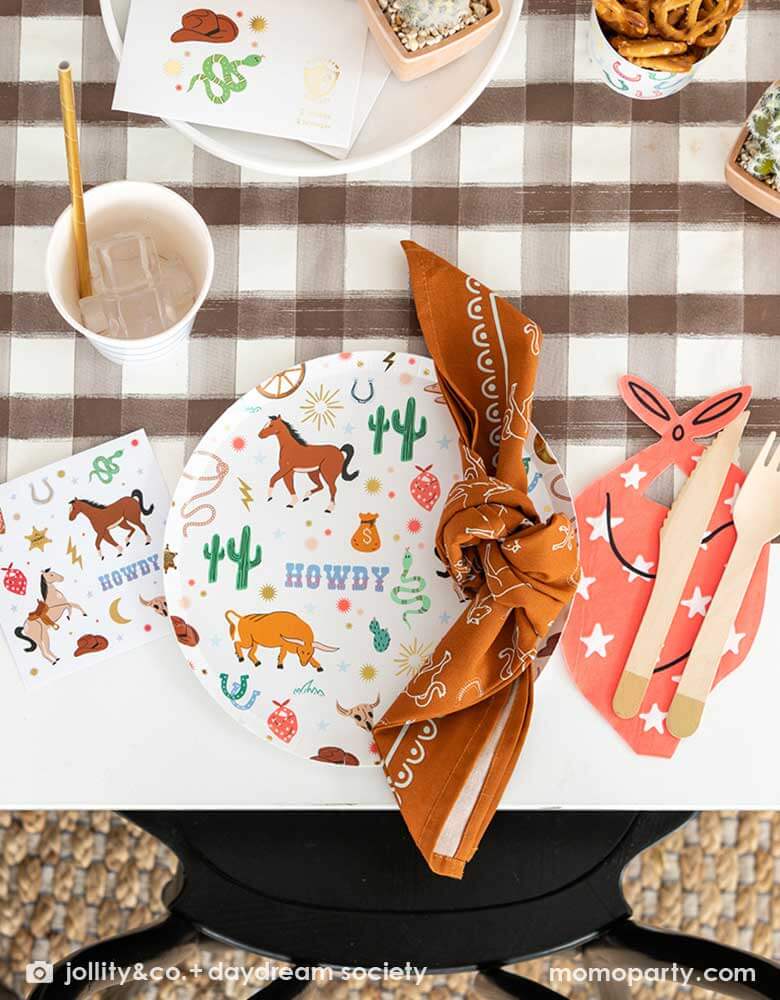 A western cowboy themed party table tablescape featuring Momo Party's cowboy themed tableware by Jollity Co. and Daydream Society including the round cowboy icon patterned round plate, the small napkin, the bandana shaped napkin, cowboy temporary tattoos all on a brown gingham table runner, making this a modern yet fun tablescape for kid's rodeo, Western or cowboy themed birthday celebration. A perfect inpso for kid's "First Rodeo" first birthday celebration.