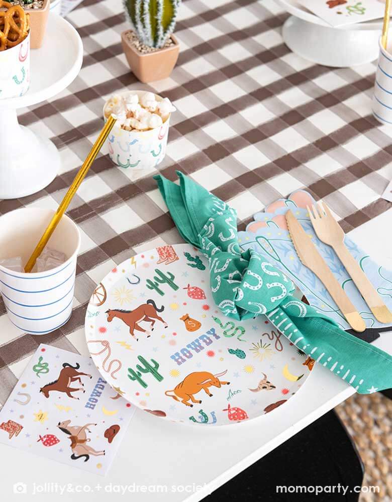 A western cowboy themed party table tablescape featuring Momo Party's cowboy themed tableware by Jollity Co. and Daydream Society including the round cowboy icon patterned round plate, the small napkin, the cowboy boot shaped napkin, horseshoe napkin all on a brown gingham table runner, making this a modern yet fun tablescape for kid's rodeo, Western or cowboy themed birthday celebration. A perfect inpso for kid's "First Rodeo" first birthday celebration.