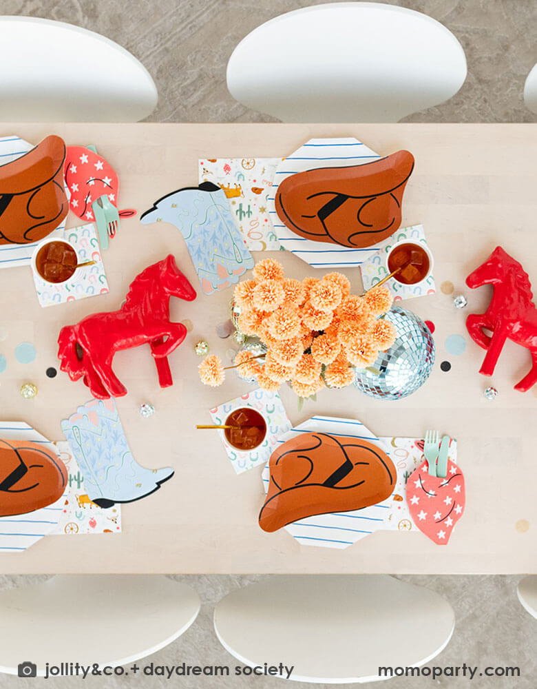 A kid's western themed party table features Momo Party's cowboy themed partyware by Daydream Society including cowboy hat shaped plates, red bandana shaped guest napkins, Western rodeo themed paper napkins and blue horseshoe small napkins. Around the tableware there are some fun disco balls around and color coordinated round confetti spread out, with rustic themed flower arrangement and red horse decorations as the centerpiece, makes this a perfect inspiration for a fun rodeo, cowboy themed celebration.